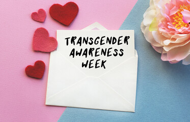 An open envelope with the text TRANSGENDER AWARENESS WEEK, on a pink and blue background with a decor of felt hearts and a flower. Flat lay, top view. LGBT and Valentine's Day concept.