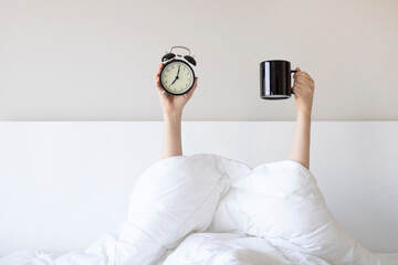 Woman showing arm raised up holding coffee cup and black alarm clock behind duvet in the bed room,...