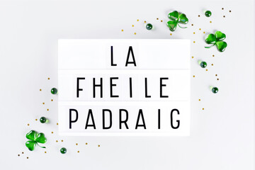 Lightbox with Saint Patricks day text written in irish, green glass shamrock symbols and gold stars confetti on white background. Flat lay Irish holiday party card Spring 17 march lucky clover design