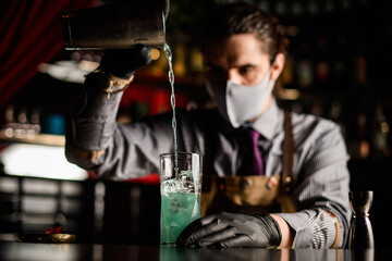 close-up of glass with ice cubes in which bartender pours blue cocktail from shaker