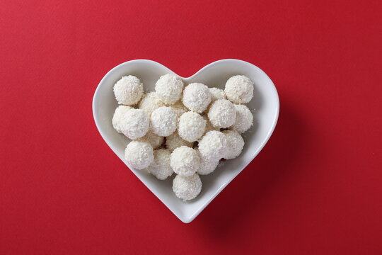 Sweet coconut rafaello candies in a heart-shaped plate on a red background. Top view