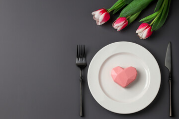 Creative table setting with hearts on a white plate, black fork and knife, tulips on a dark background. Valentine's Day, Wedding Day, Birthday, Women's Day and Mother's Day. Flat lay. Copy space