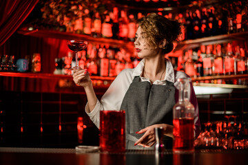beautiful woman bartender at bar and holds glass of cocktail and looks at it