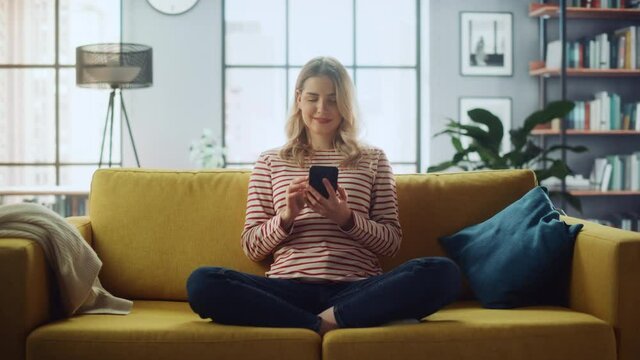 Beautiful Caucasian Female Using Smartphone in Stylish Living Room while Sitting on a Cozy Couch Sofa. Young Woman Resting at Home, Browsing Internet, Using Social Networks, Having Fun in Flat.
