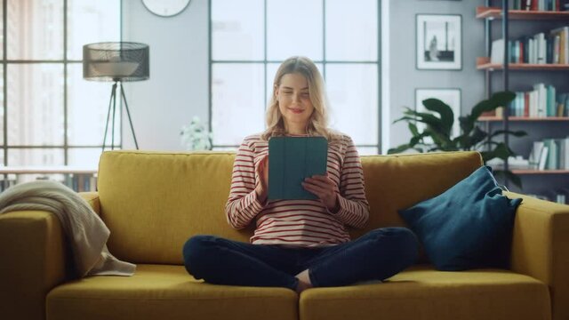 Beautiful Caucasian Female Using Tablet Computer in Stylish Living Room while Sitting on a Cozy Couch Sofa. Young Woman Resting at Home, Browsing Internet, Using Social Networks, Having Fun in Flat.