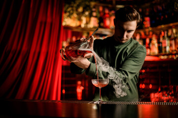 Fototapeta na wymiar young bartender pouring bright steaming cocktail into glass on bar counter