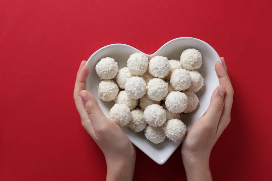 Sweet coconut rafaello candies in a heart-shaped plate in female hands on a red background. View from above