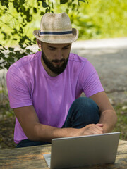 A young bearded guy in jeans, a pink t-shirt and a hat sits behind a wooden one. A freelancer looks at the screen of his laptop and thinks about something. The concept of travel, business.