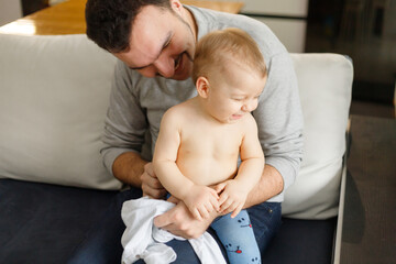 Father dresses infant in living room sitting on couch at home. Clothes baby undershirts and sliders on baby close-up. Home care and baby care concept.