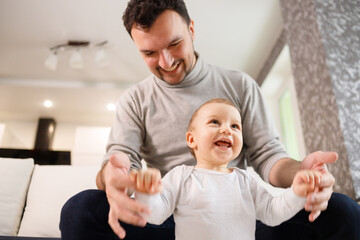 Young father assisting walking his infant son teaching to walk at home. Daddy helps son attempting to walk