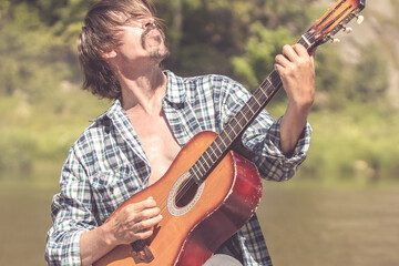 Rock musician man in an open shirt enthusiastically plays the old guitar in nature. Summer, clear...
