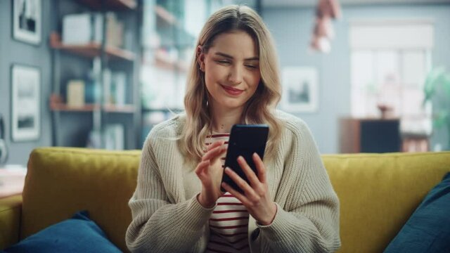 Beautiful Caucasian Female Using Smartphone in Stylish Living Room while Resting on a Cozy Couch Sofa. Young Woman at Home, Browsing Internet, Using Social Networks, Having Fun in Flat. Zoom In Shot.