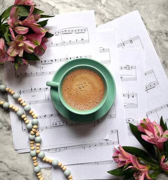 A Couple Of Sheet Music Thrown On The Table With A Cup Of Coffee On Top , Pink Flowers And Wood Beads Garland Shot On A Marble Surface. Musical Background.  Top View, From Above, Flat Lay.