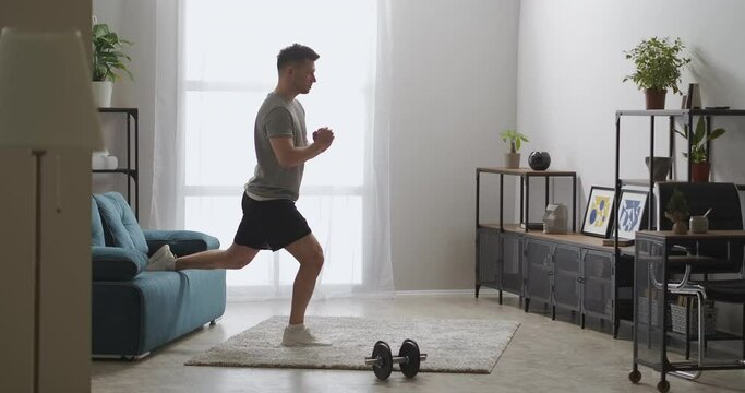 home fitness training of adult man in apartment, athletic guy is squatting in room at morning, healthy lifestyle and workout