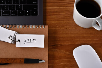 On the desk there is a laptop, a cup of coffee, and a word book with the word STEM written on it. It was an abbreviation for Science, Technology, Engineering and Mathematics.