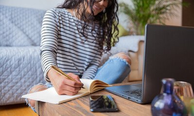 Close up of a happy young beautiful girl is sitting and writing on her notebook. Behind her is a...
