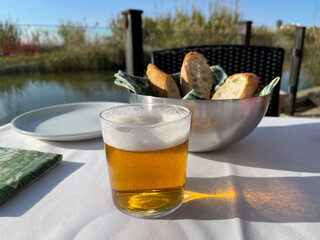 Close-up shot of a table in outdoor cafe with a glass of beer and bowl with fresh bread. Rural scenery in background. El Palmar, Spain