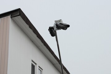 one black surveillance camera on a long metal tube against the background of the white wall of the house and the gray sky