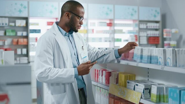 Pharmacy: Portrait of Professional Black Pharmacist Uses Digital Tablet Computer, Checks Inventory of Medicine, Drugs, Vitamins on a Shelf. Drugstore Store Arranging Health Care Products. Zoom in Shot