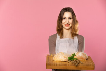 An attractive young saleswoman holds two plump breads decorated with herbs and ears of ripe grain on a wooden board. Seamless light pink background.