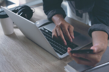 Business man hand using mobile phone and online working on laptop computer on table, close up