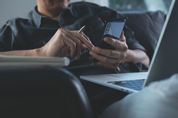 Casual business man sitting on sofa, using mobile phone surfing the internet and online working on laptop computer from home