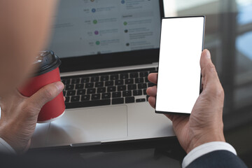 Mockup image, business man hand holding blank screen mobile smart phone and cup of coffee with laptop computer on table at office