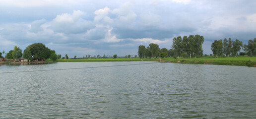 Beautiful scene of a natural rural landscape with a pond, crop fields, trees and clouds in sky.
