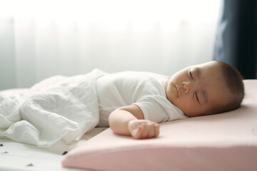 Asian little boy lying down on a bed. A cute infant sleepy on a pink pillow, white blanket in...