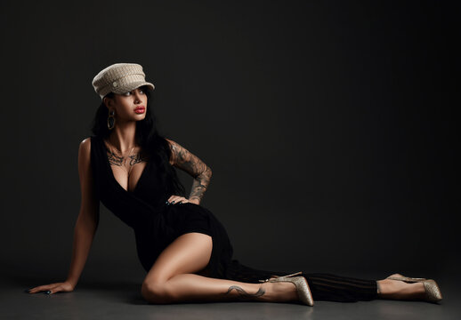 Fashionable woman with pouty lips and tattoo on body in evening pinstripe gown with deep neckline is lying on floor looking at copy space over dark background. Stylish look, fashion, glamour concept