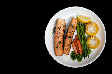 Two pieces salmon steak with vegetables, grilled corn, asparagus, carrots, green peas, lemon, pepper on isolated black background with clipping path. Top view.