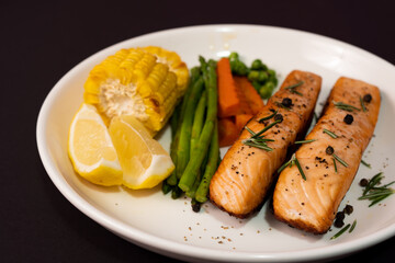 Two pieces salmon steak with vegetables, grilled corn, asparagus, carrots, green peas, lemon, pepper on black background.