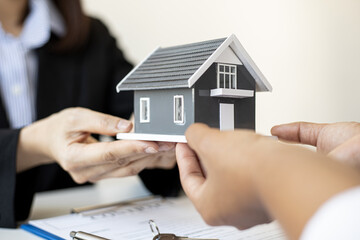 Close-up female home salesman handing over a gray small house model with two hands to the customer...