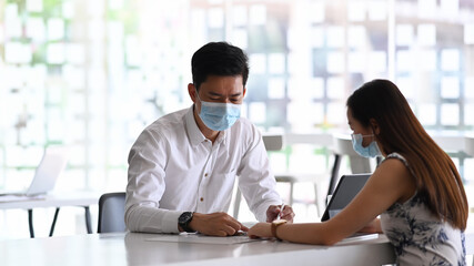 Two young business people in protective mask working together on laptop computer and analyzing report at office desk.