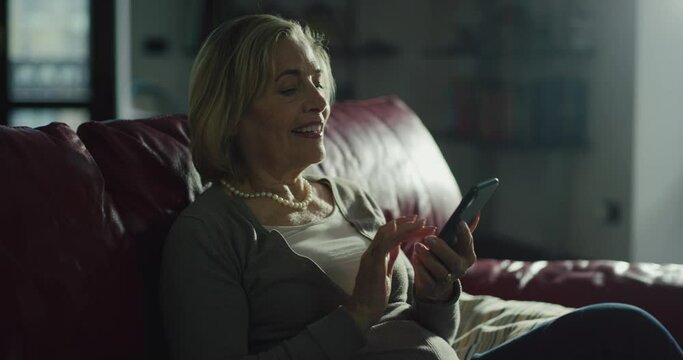 Cinematic shot of happy mature senior woman using smart phone for send messages and emails, make calls and use apps while sitting on sofa at home. Concept of technology, modern generation, connection.