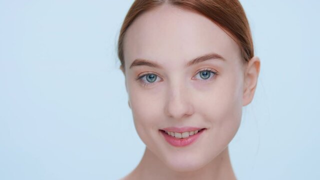 Close-up beauty portrait of young attractive ginger woman softly touches her skin running fingers from her hand up to the shoulder, looks at camera and smiles