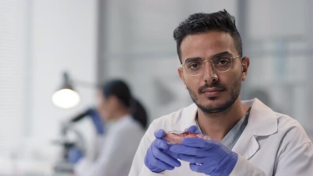 Close up slowmo portrait of middle eastern researcher examining artificial meat sample in white laboratory looking at camera