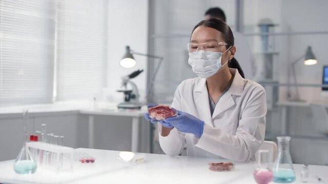 Medium slow-motion portrait of Asian female microbiologist in safety mask and glasses sitting at lab table, examining lab-grown meat samples and looking at camera