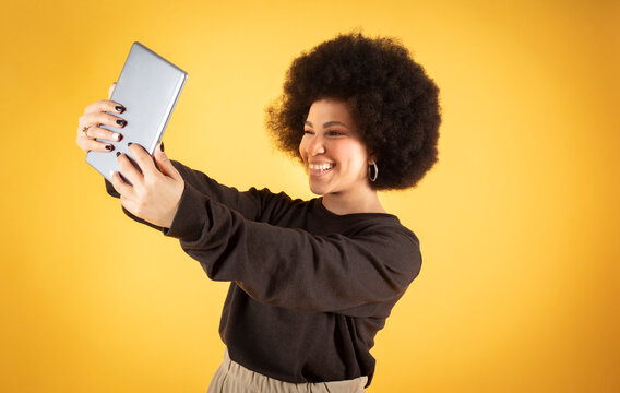 Mixed afro hair woman in casual wear, happy, makes selfie with tablet, using tablet, communications and social media, yellow background, copy space