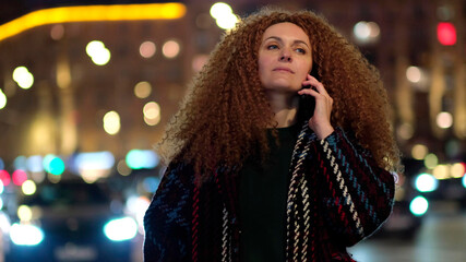 pretty woman in trendy coat walking on metropolis street with night lights and talking on the phone.