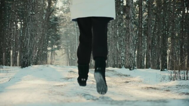 Young woman with long loose hair in warm white jacket runs along snowy road in sunny winter forest backside view slow motion