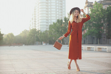 Beautiful young woman in stylish red dress and hat with handbag on city street