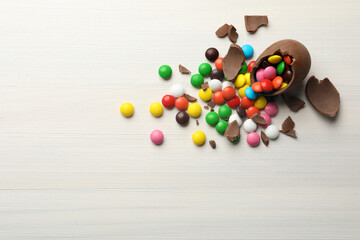 Broken chocolate egg and colorful candies on white wooden table, flat lay. Space for text