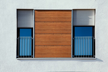 two windows side by side next to each other on white wall of an apartment building with european grey shutters half way down to block sun