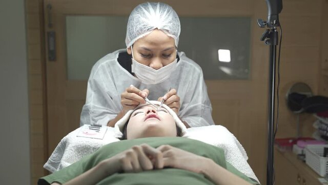 asian makeup artist making lashes salon procedure. Woman's lashes extension. Professional wearing medical face mask hat and PPE suite for hygienic service during covid-19. Young, healthy skin.