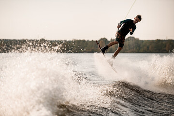 Young man wakeboarder holds cable and actively rides on splashing wave.