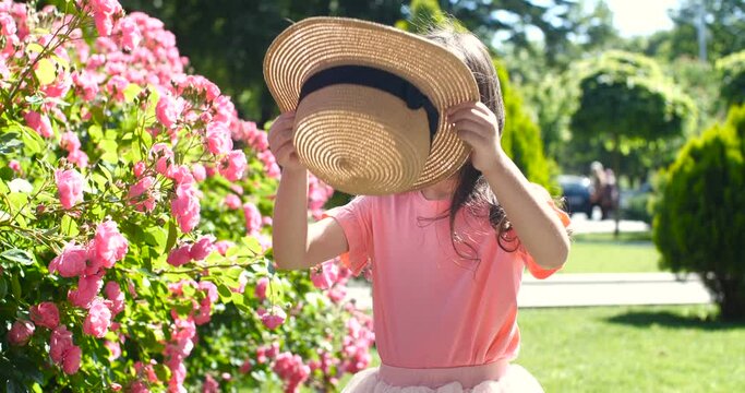 Pretty Caucasian girl of preschool age with long brown hair in pink dress putting on straw hat and joyfully  dancing  near rose bushes  in a park on sunny day. 4k 50 fps slow motion
