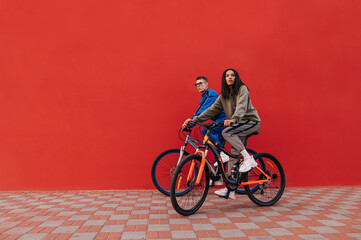 Fototapeta na wymiar Photo of stylish man and woman riding a bicycle on the street on a background of a red wall. Stylish sports couple of cyclists walking down the street on the weekend