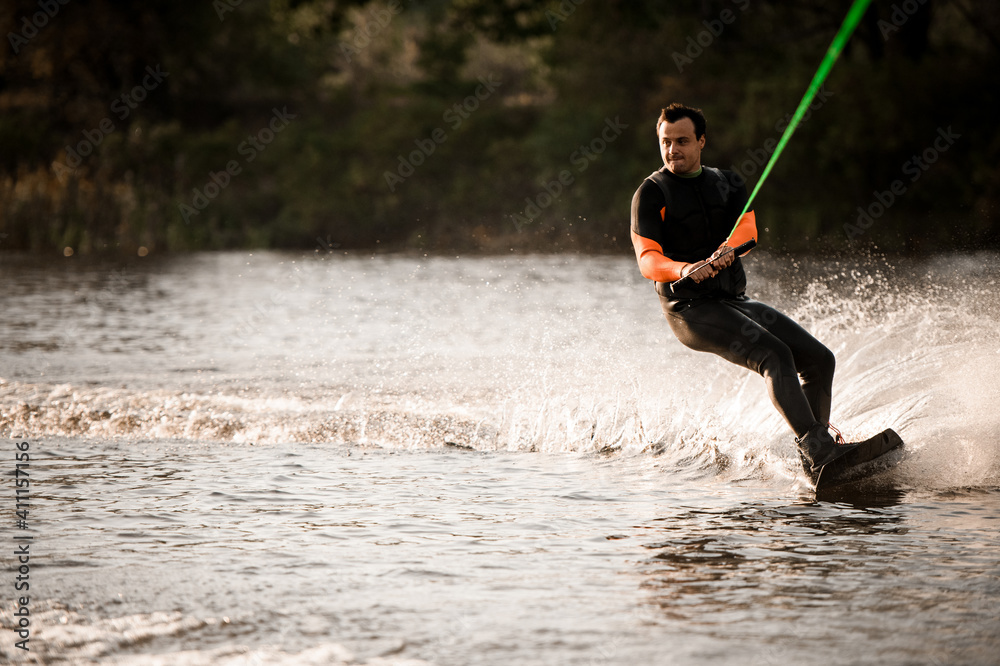 Wall mural adult man in wetsuit holds cable in his hands and balancing on splashing wave