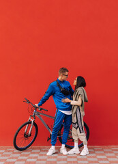Vertical photo of two sports lovers on a background of a red wall standing with a bicycle, hugging and looking at each other. Copy space